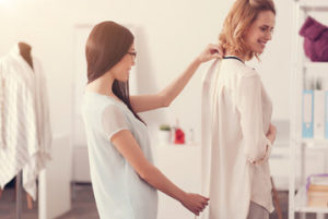 due donne in atelieur una prende le misure alla schiena dell'altra per confezionare un'abito su misura sullo sfondo manichino e appendiabiti 
